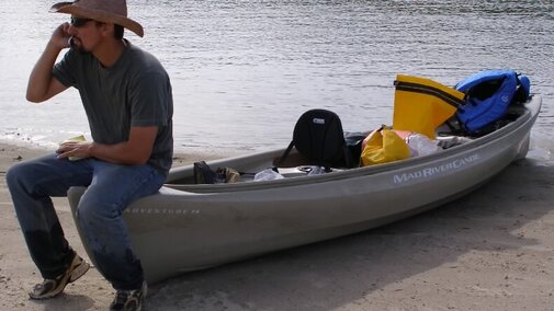 Joel Jorgensen on the phone on a canoe on a river sandbar